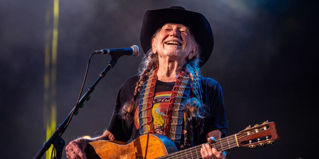 A smiling, long-haired musician in a cowboy hat playing an acoustic guitar on stage.