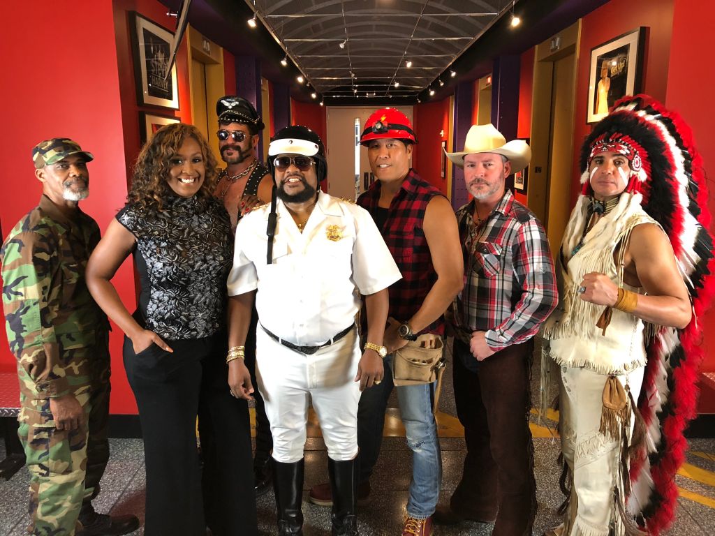 A group of people dressed in various costumes and uniforms, including military, police, and Native American-inspired attire, posing together in a hallway with red walls and framed artwork.